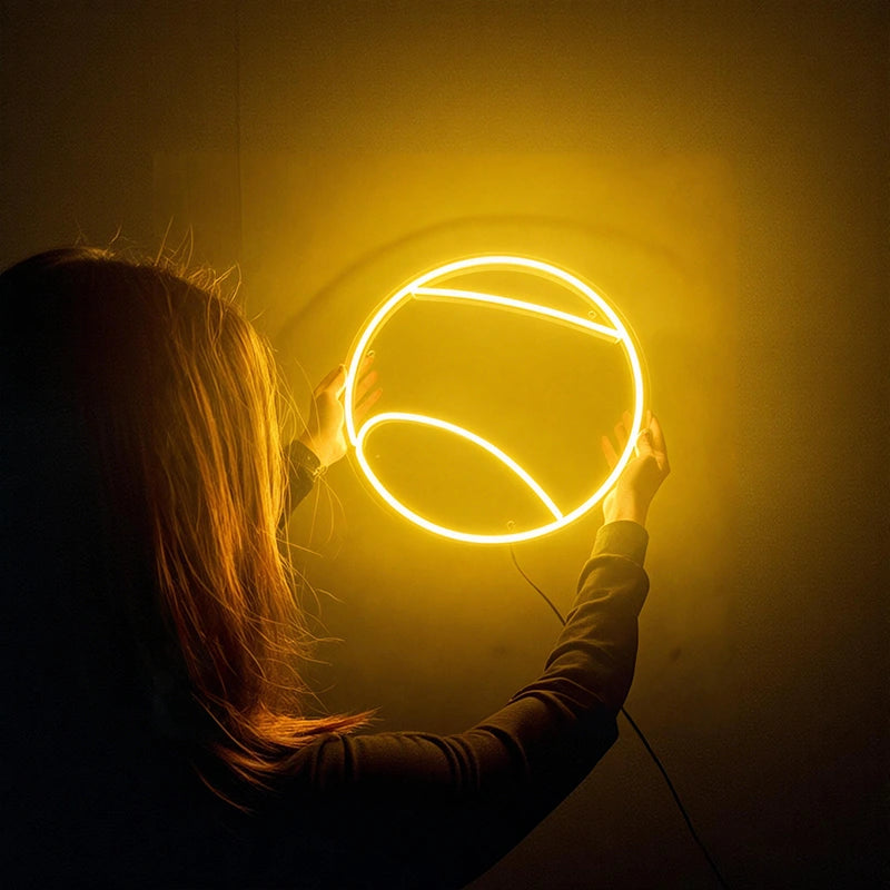 Person holding a round lemon yellow tennis ball neon light against a dark background