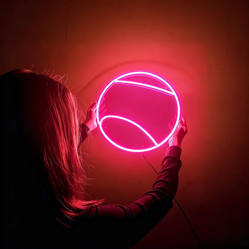 Person holding a round hot pink tennis ball neon light against a dark background