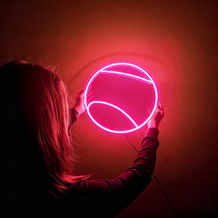 Person holding a round hot pink tennis ball neon light against a dark background