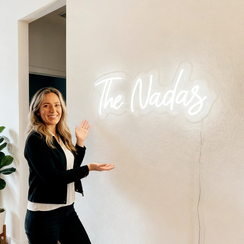 A smiling woman stands indoors beside a glowing white neon-style wall sign that reads 'The Nadas' on a light beige wall.