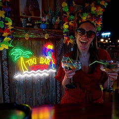 Woman holding two cocktails in front of a Tiki Bar Open neon sign.