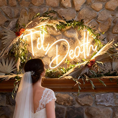 Neon sign with 'Til Death' surrounded by decorative plants and a stone wall.