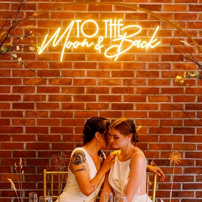 Two women in white dresses sit closely together, sharing an intimate moment at a table. A neon sign reading 'TO THE Moon & Back' glows above them against a red brick wall, and minimal floral decor surrounds them.