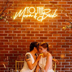 Two women in white dresses sit closely together, sharing an intimate moment at a table. A neon sign reading 'TO THE Moon & Back' glows above them against a red brick wall, and minimal floral decor surrounds them.