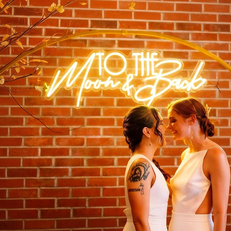 Two women standing in front of a neon sign that reads 'To the Moon & Back' against a brick wall.