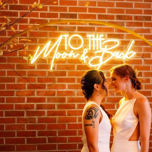 Two women standing in front of a neon sign that reads 'To the Moon & Back' against a brick wall.