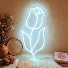 The neon white light of a tulip-shaped sign falls on a wall with flowers and books placed beside it.