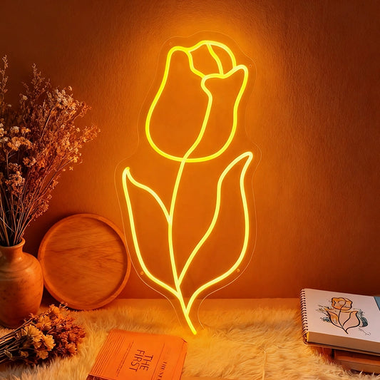 The neon orange light of a tulip-shaped sign falls on a wall with flowers and books placed beside it.