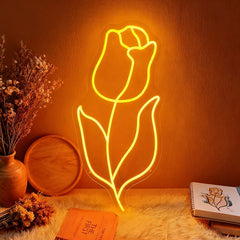 The neon orange light of a tulip-shaped sign falls on a wall with flowers and books placed beside it.