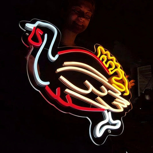 A large illuminated neon sign shaped like a stylized turkey, glowing with red, lemon yellow, white, and warm white tubing against a dark background. A person is faintly visible behind the sign.