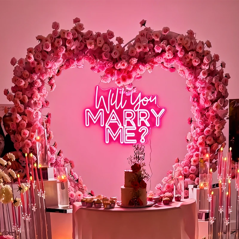 Neon sign 'Will you marry me?' surrounded by pink floral arch and cake on a table.