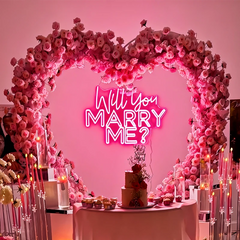 Neon sign 'Will you marry me?' surrounded by pink floral arch and cake on a table.