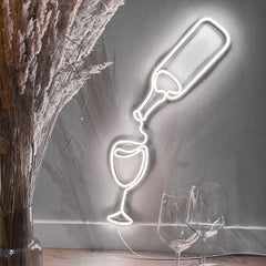 Neon light shaped like a champagne bottle and glass on a gray wall with two real wine glasses and a vase.