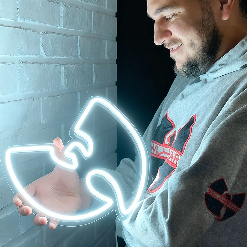 Man holding a white neon sign shaped like a Wu-Tang Clan logo in front of a brick wall.