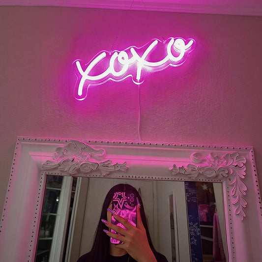 A hot pink neon sign spelling 'xo xo' glowing above a large ornate white mirror, with a person taking a mirror selfie in a room.