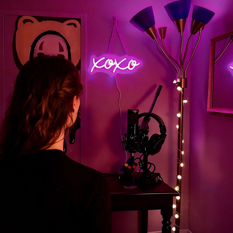 A person viewed from behind in a dimly lit room with hot pink ambient lighting, facing a desk setup with a neon 'XOXO' sign, headphones, and a microphone. A floor lamp stands to the right against a dark wall.