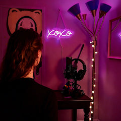 A person viewed from behind in a dimly lit room with hot pink ambient lighting, facing a desk setup with a neon 'XOXO' sign, headphones, and a microphone. A floor lamp stands to the right against a dark wall.