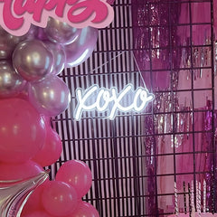 Pink and silver balloon garland, a black wire grid backdrop, and a glowing white 'xo xo' neon sign against a pink tinsel background.