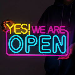 Neon sign that reads 'YES! WE ARE OPEN' held by a person against a dark background