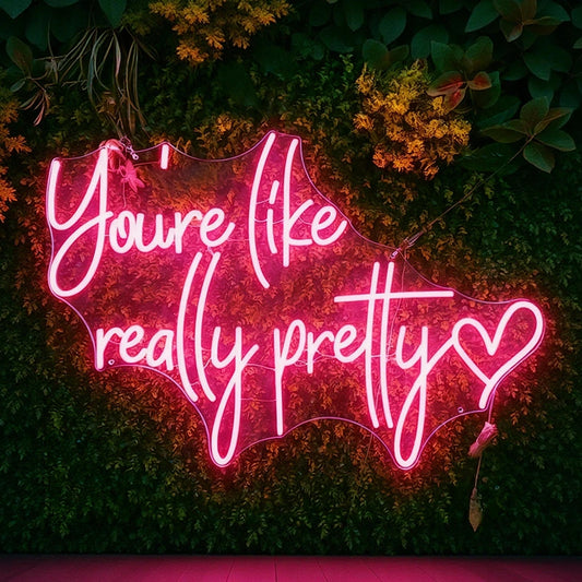 You're Like Really Pretty hot pink neon sign mounted on a green foliage wall, perfect for bedroom, beauty salon, selfie wall, wedding, and event decor.