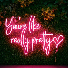 You're Like Really Pretty hot pink neon sign mounted on a green foliage wall, perfect for bedroom, beauty salon, selfie wall, wedding, and event decor.