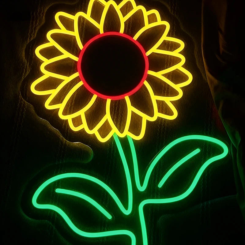 Neon light sculpture of a sunflower with lemon yellow petals, red center, and green stem on a dark background.