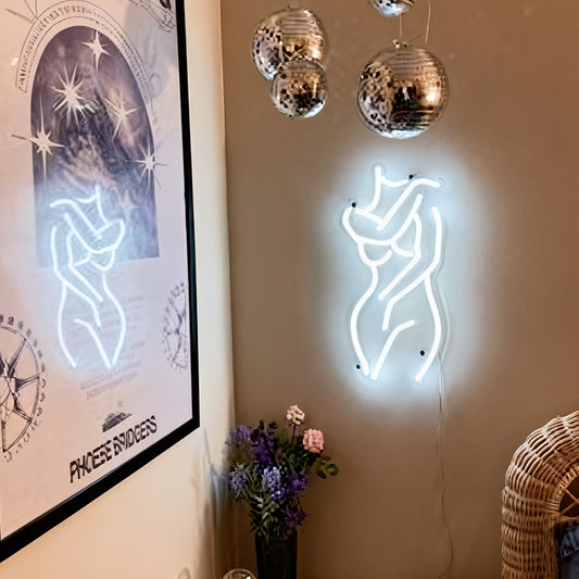 Neon sign of a female body on a wall with a framed poster and decorative lights.