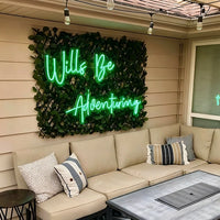 A green neon sign reading 'Wills Be Adventuring' mounted on a faux greenery wall above a beige outdoor sectional sofa with decorative pillows, in a patio setting.