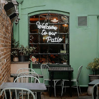 A green-painted building exterior with an arched window displaying the words 'Welcome to our Patio' in white script. Several outdoor dining tables and chairs are arranged in front of the window, with potted plants and a brick wall visible on the left.