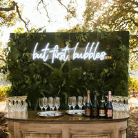Outdoor event setup with a wooden bar counter, champagne bottles, and many wine glasses in front of a green leaf wall with a glowing white neon sign that reads 'but first bubbles'.
