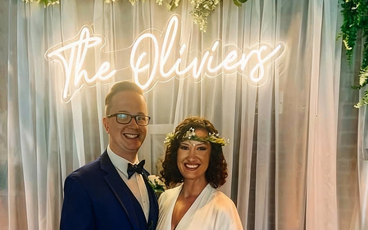 Smiling bride and groom standing beneath a warm white custom “The Oliviers” LED neon sign at a wedding reception with elegant draped curtains and greenery décor.