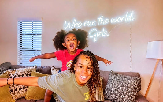 Mother and daughter posing in a living room with a glowing white 'Who Run the World Girls!' LED neon sign on the wall, creating a fun and empowering atmosphere.